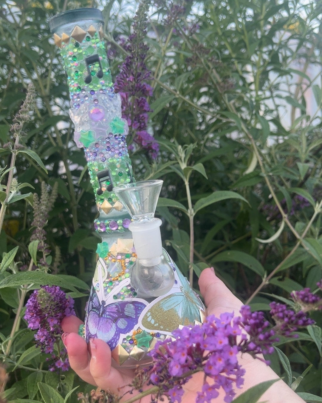 A hand holding a decorated glass bong surrounded by green leaves and purple flowers outdoors.